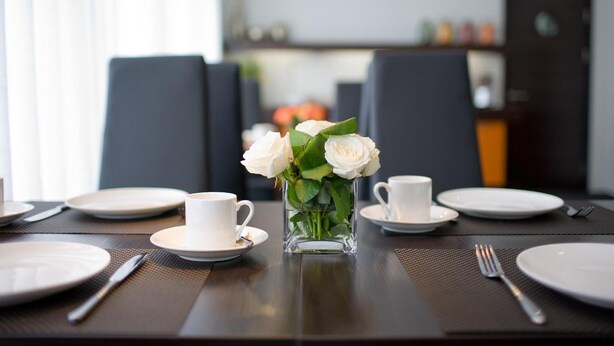 Une table à manger dressée avec de l’assiette blanche, des tasses et un vase transparent contenant des fleurs blanches.