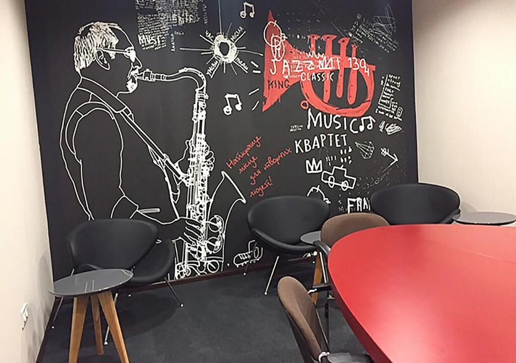 A room with a blackboard featuring musical notes and a saxophone drawing, a red table, and black chairs.