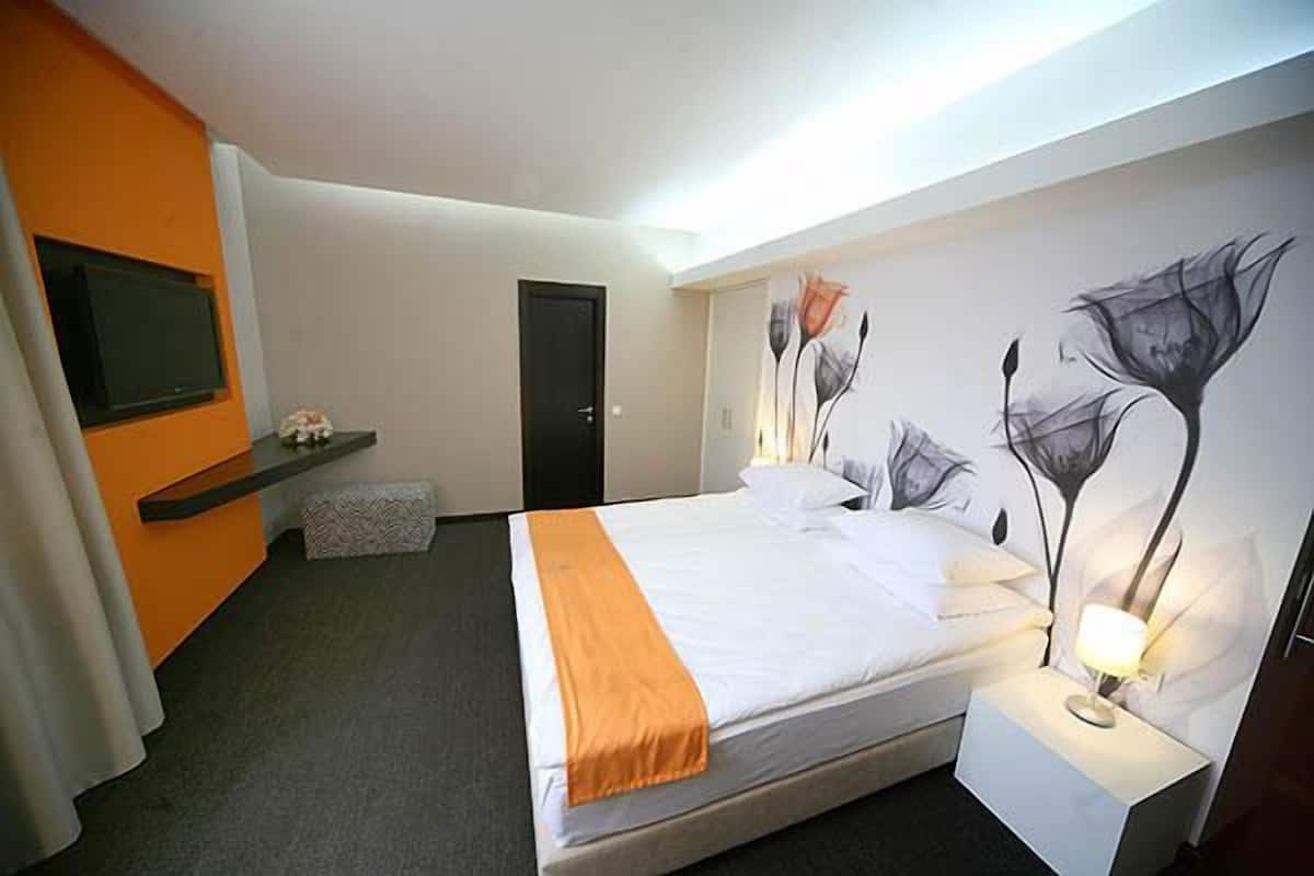 A hotel room with a bed, a nightstand with a lamp, a TV mounted on the wall, and a painting of flowers on the wall.