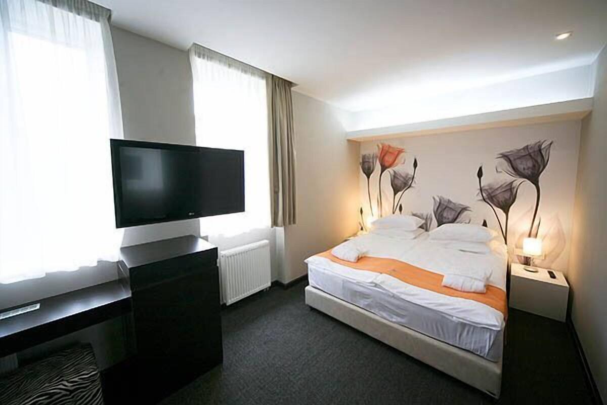 A hotel room with a bed, a TV, a desk, and a painting on the wall.