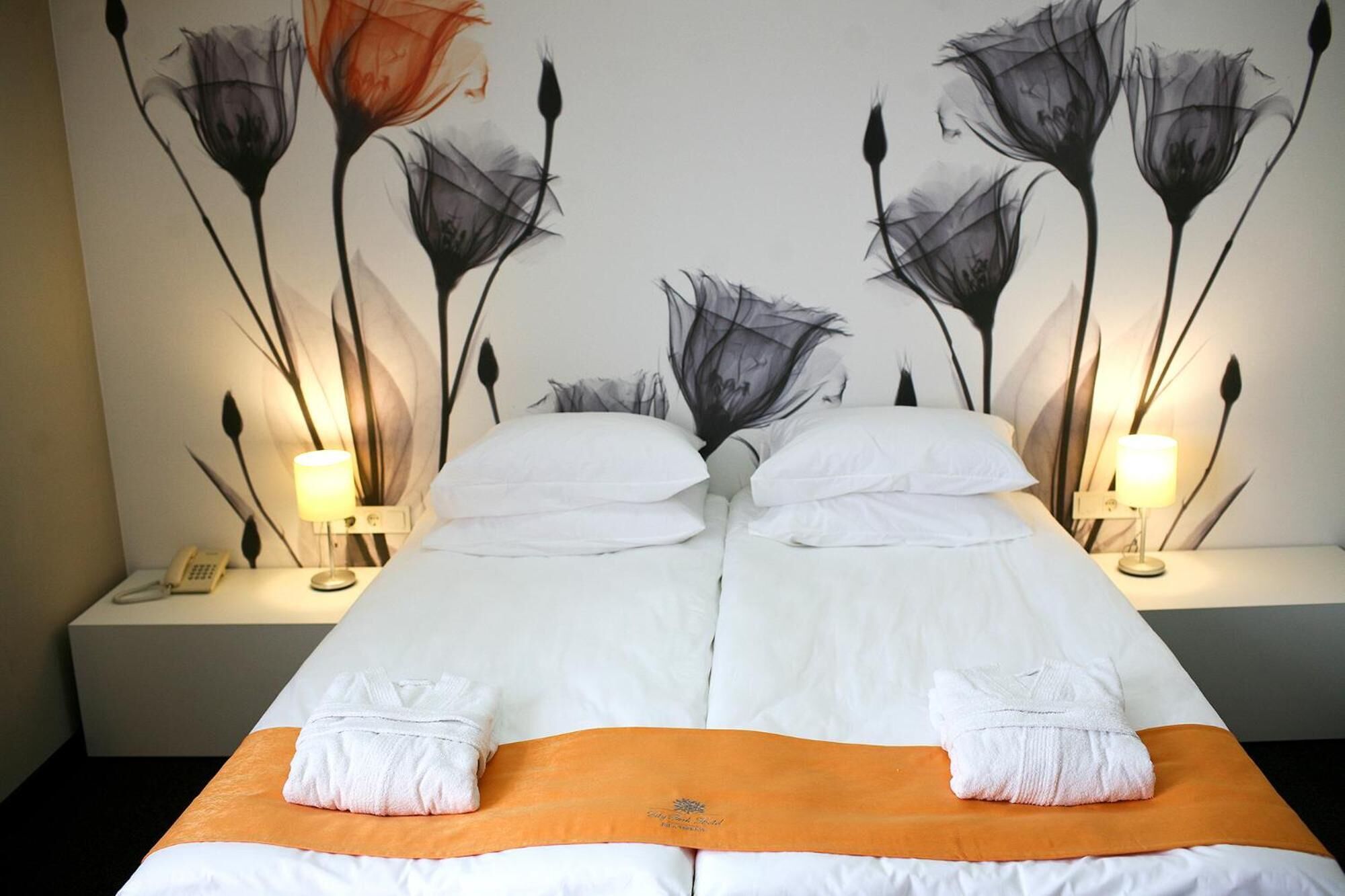 Ein Doppelbett mit weißen Bettwäsche, zwei weiße Handtücher und ein orangefarbener Tagesdecke. Zwei Nachttische mit Lampen und ein Telefon sind vorhanden. An der Wand hängt ein großes Blumenbild.
