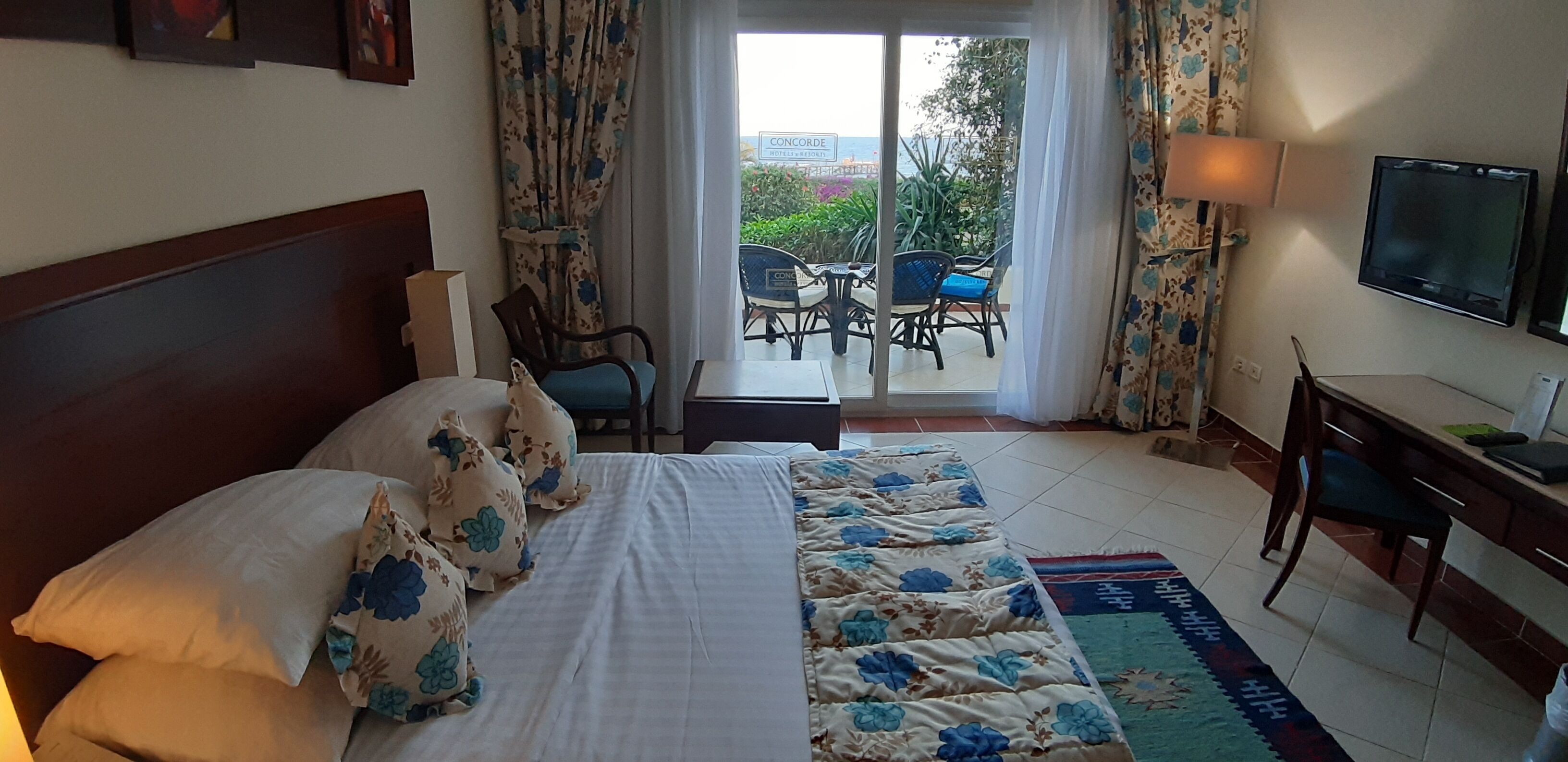A hotel room with a bed, a desk, a TV, and a balcony with chairs and a table.