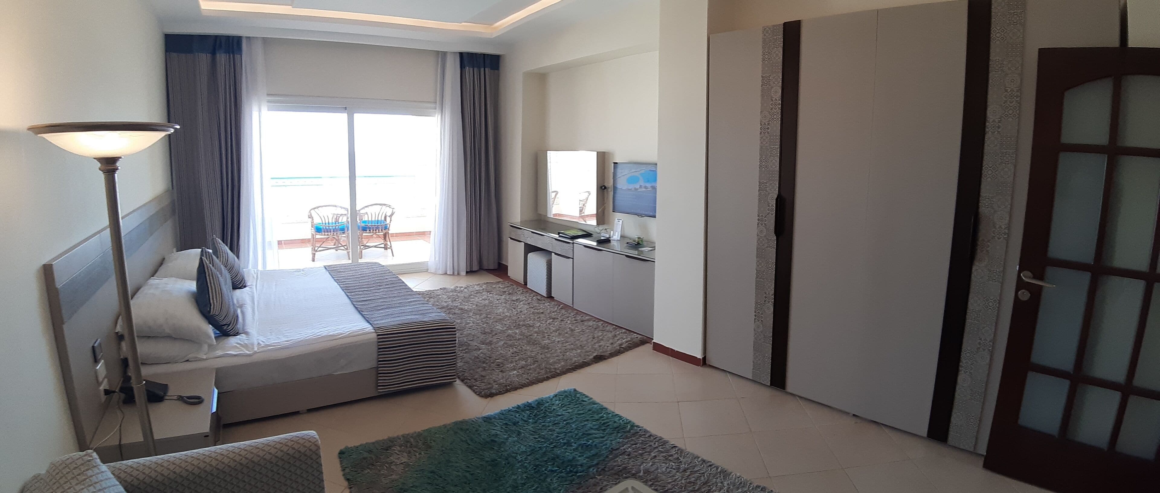 A hotel room with a bed, bedside lamp, a small kitchenette, a TV, and a sliding door.