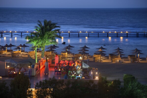A beachside resort with a pier extending into the water, lounge chairs, umbrellas, and a palm tree.