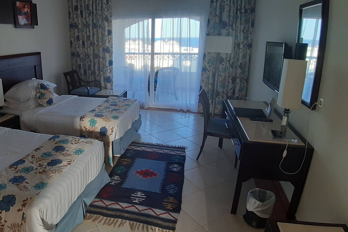 A hotel room with two beds, a desk, a TV, and a view of the beach through the window.