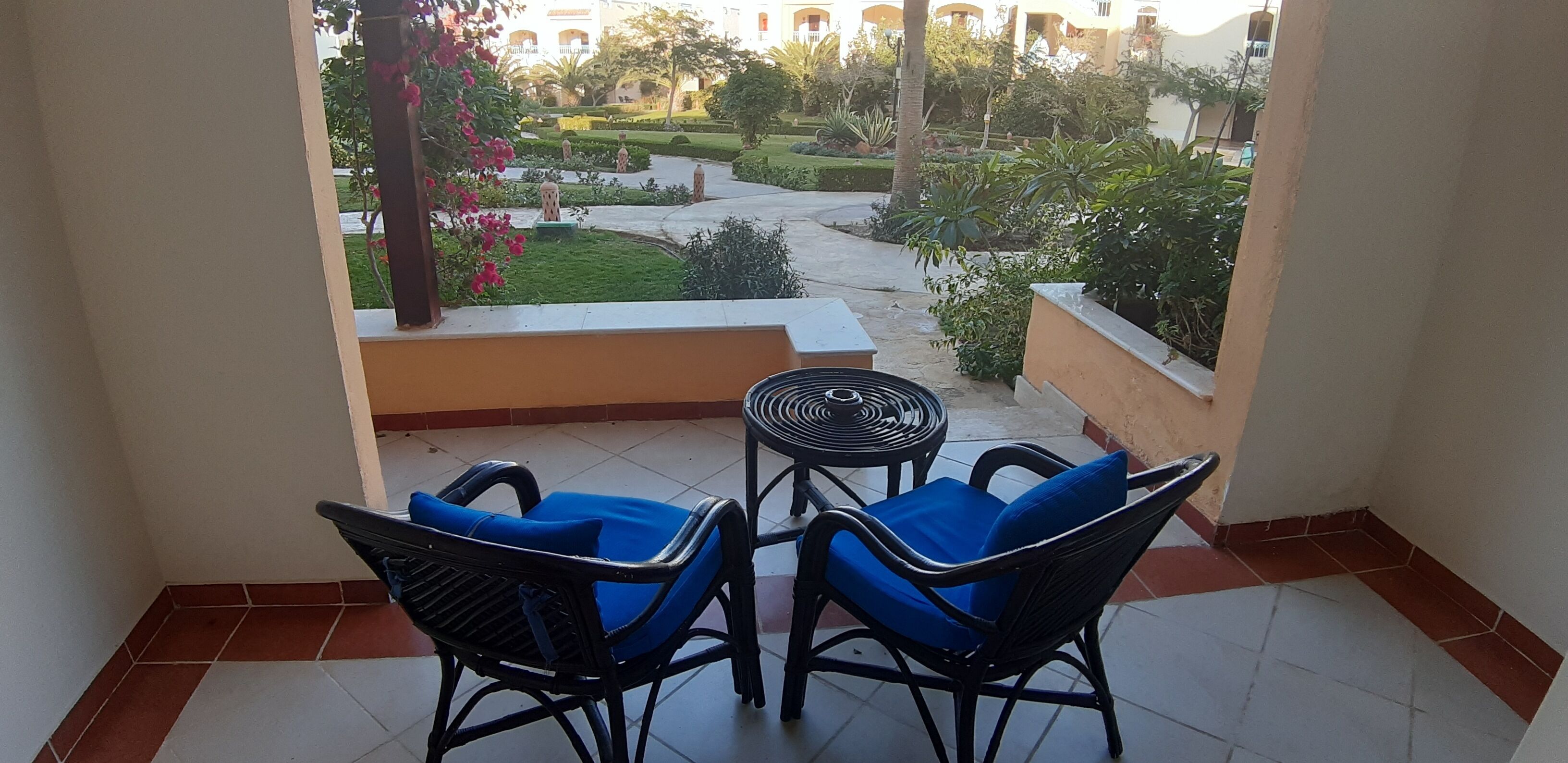 A balcony with two blue cushioned chairs and a small round table, overlooking a well-maintained garden with pathways and various plants.