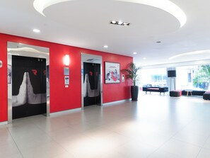 A modern lobby with red walls, two elevators, a large potted plant, and a seating area with a view of the street. - Red Planet Makati Avenue Manila (Makati)