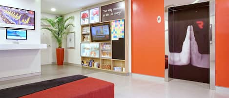 Un espace d’accueil comprenant un banc rouge, une plante en pot, un téléviseur fixé au mur et une bibliothèque contenant divers objets.