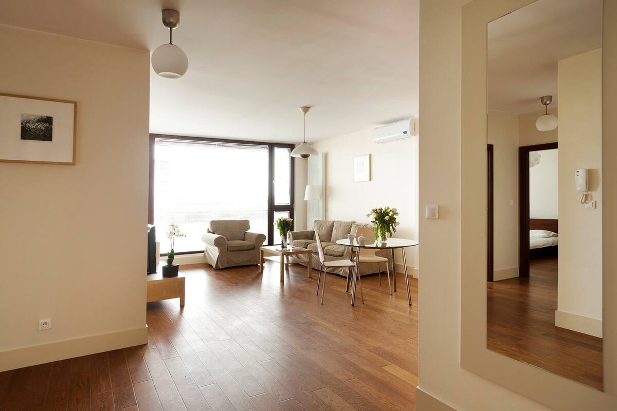 Un salon spacieux doté d’un parquet, d’un ensemble de canapés, d’une table à manger avec des chaises et d’une grande fenêtre laissant entrer la lumière naturelle.