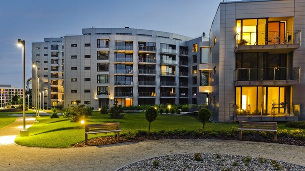Un immeuble moderne à plusieurs étages, doté de balcons, d’une pelouse bien entretenue et d’un banc.