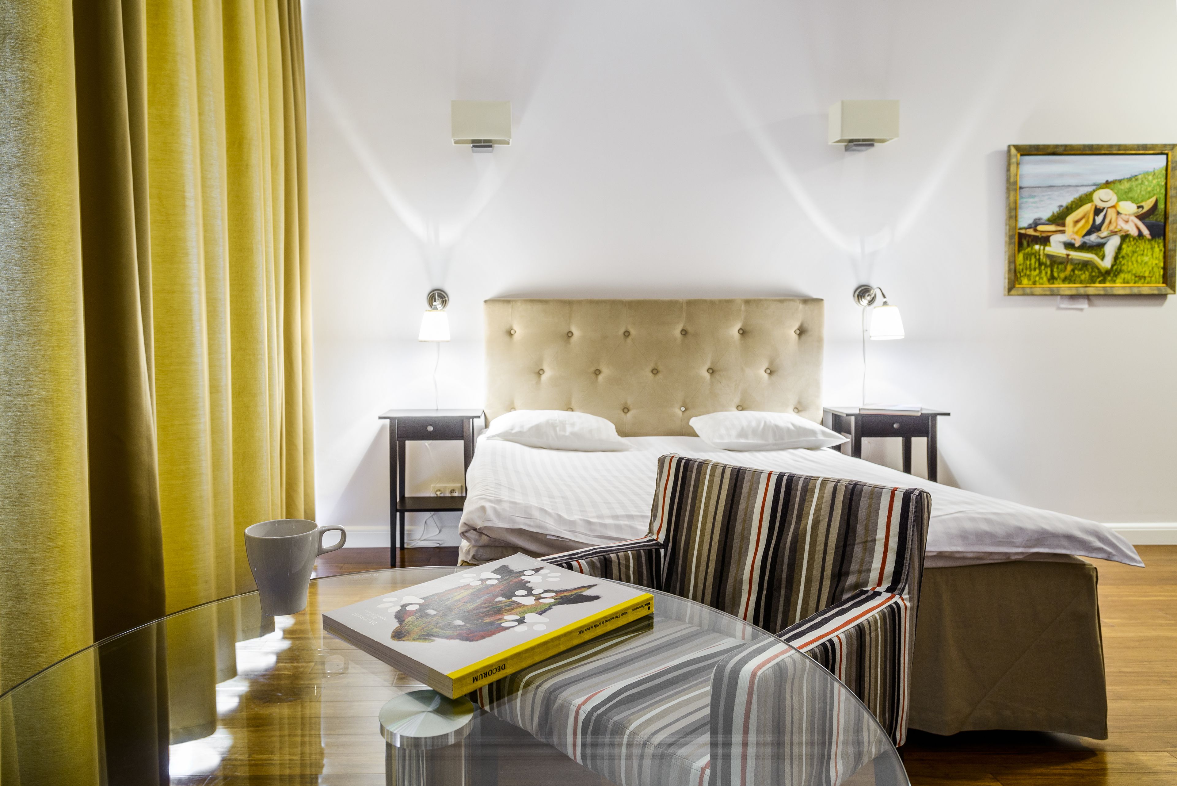 Ein Hotelzimmer mit einem Bett, Nachttischen, einem Glastisch, einem gestreiften Sessel, einem Bild an der Wand und einem Fenster mit Vorhängen.
