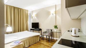 A hotel room with a bed, a desk, a chair, a TV, a kettle, and a small table with a plant.