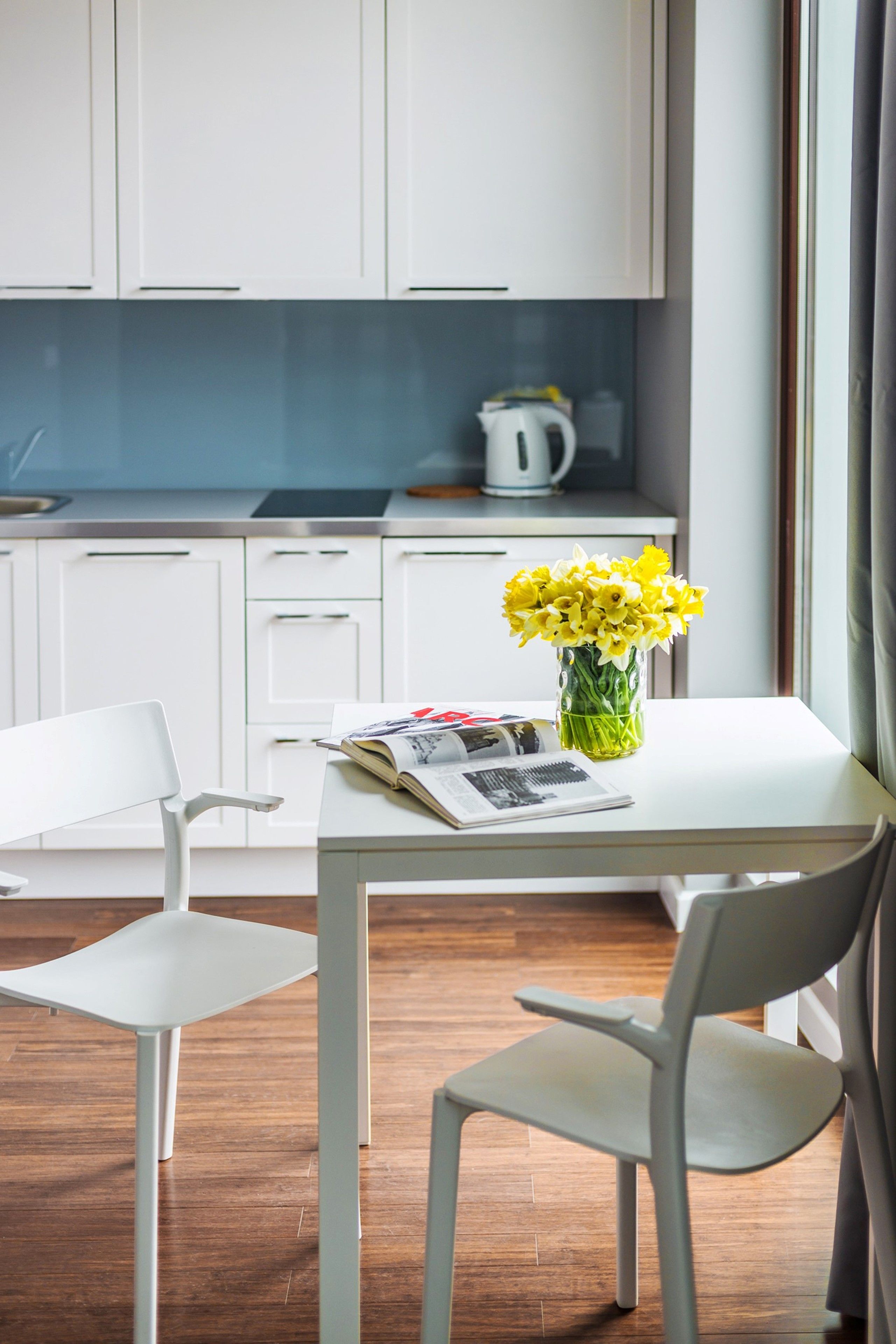 A modern kitchen with white cabinets, a white table with a vase of yellow flowers, and a kettle on the counter.