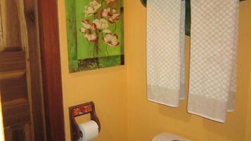 Casa de banho com sanita, suporte para papel higiénico, toalheiro com duas toalhas e um quadro decorativo de uma flor na parede.