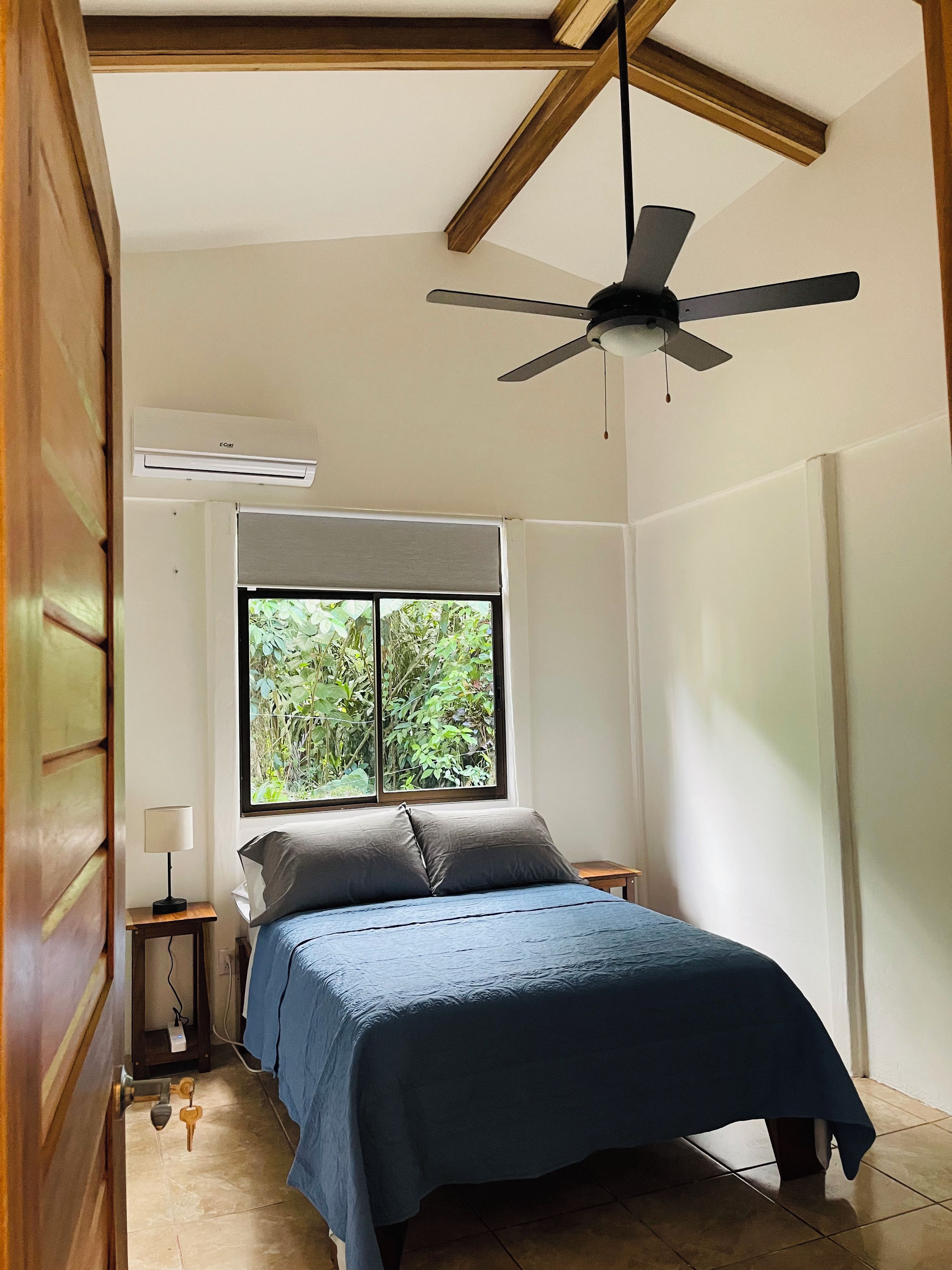 Un dormitorio con cama, ventilador de techo, ventana con vista a follaje y una mesita de noche con lámpara.