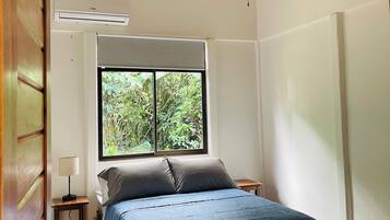 Un dormitorio con cama, ventilador de techo, ventana con vista a follaje y una mesita de noche con lámpara.