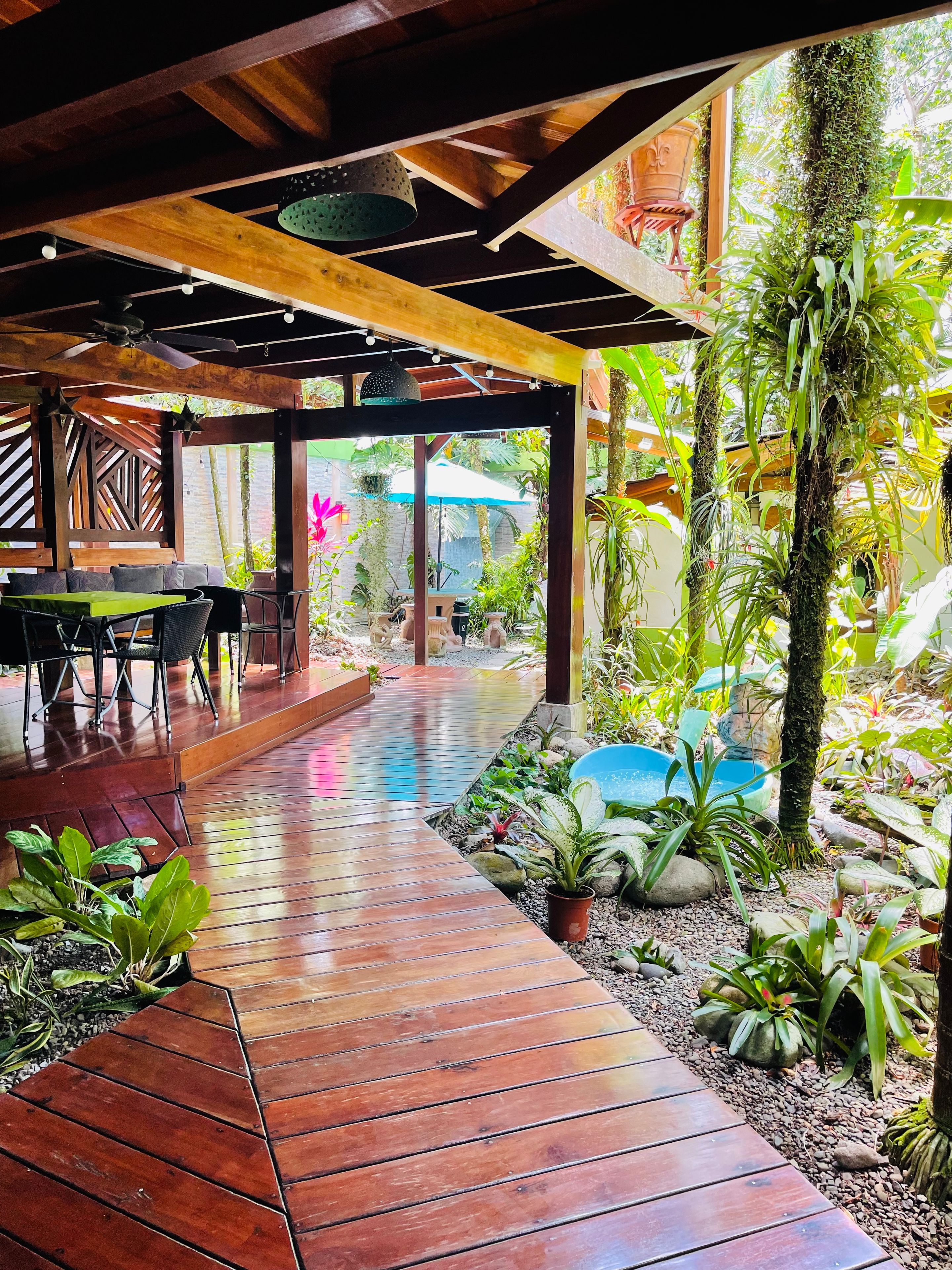 Una terraza exterior de madera con una mesa y sillas, rodeada de diversas plantas y una pequeña fuente de agua.