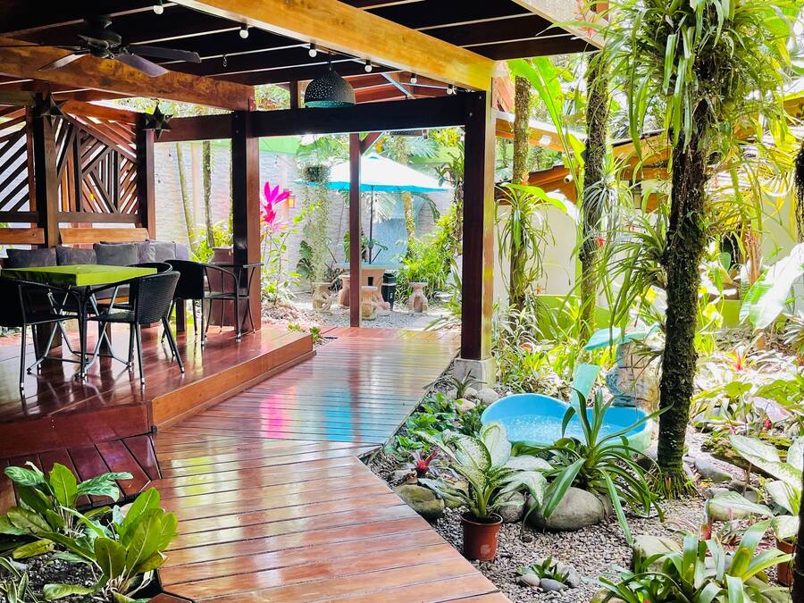Una terraza exterior de madera con una mesa y sillas, rodeada de diversas plantas y una pequeña fuente de agua.