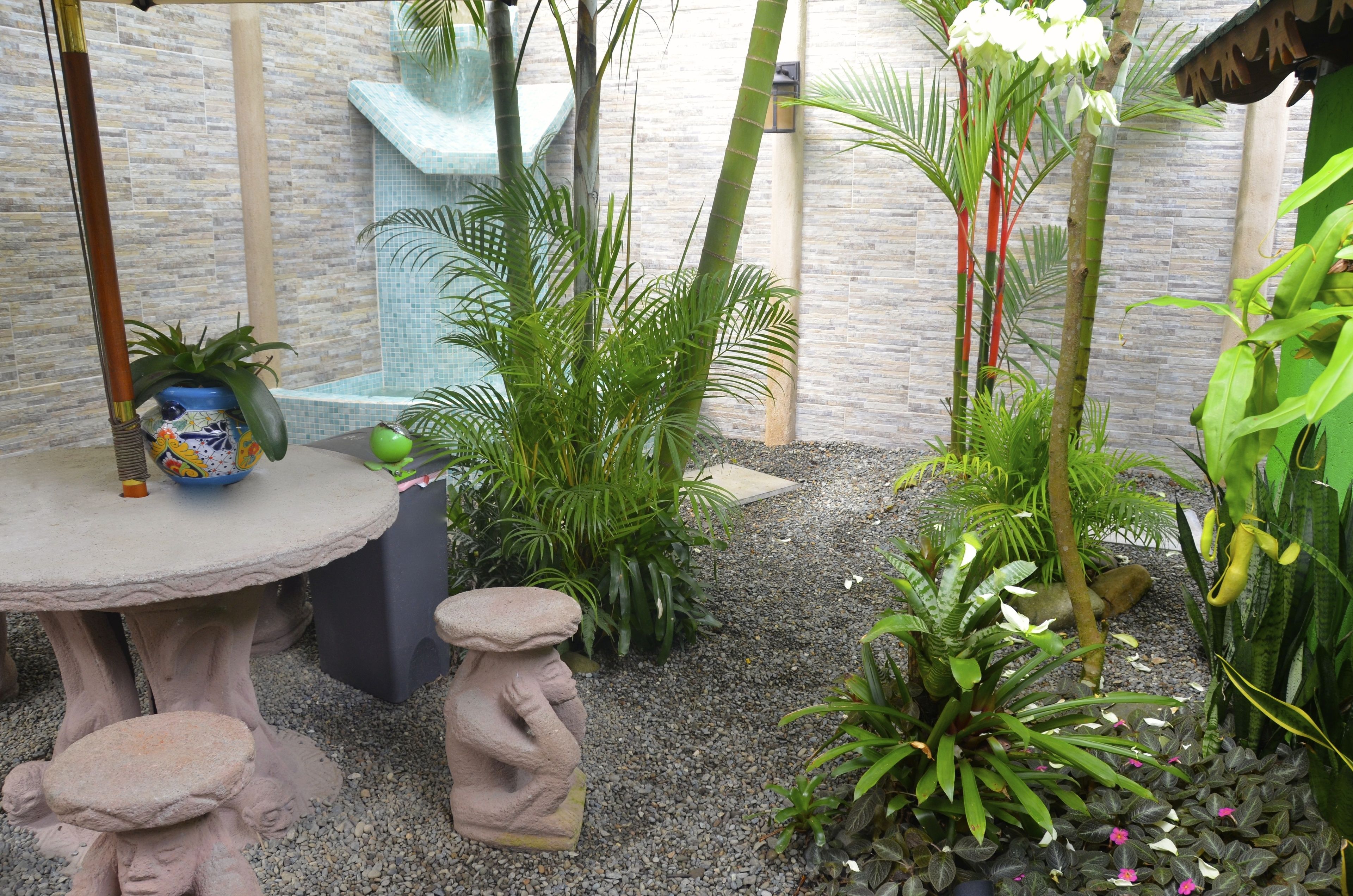 Ein Gartenbereich mit Steintisch, zwei Stehstühlen und verschiedenen Pflanzen, darunter Palmen und Orchideen.