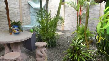 Ein Gartenbereich mit Steintisch, zwei Stehstühlen und verschiedenen Pflanzen, darunter Palmen und Orchideen.