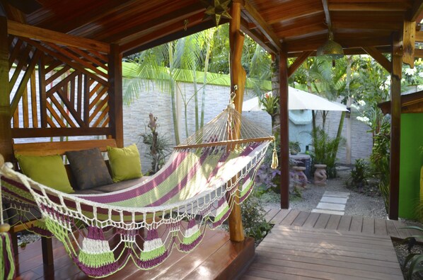 A hammock under a wooden pergola, a wooden bench with cushions, and a garden with plants and a stone wall.