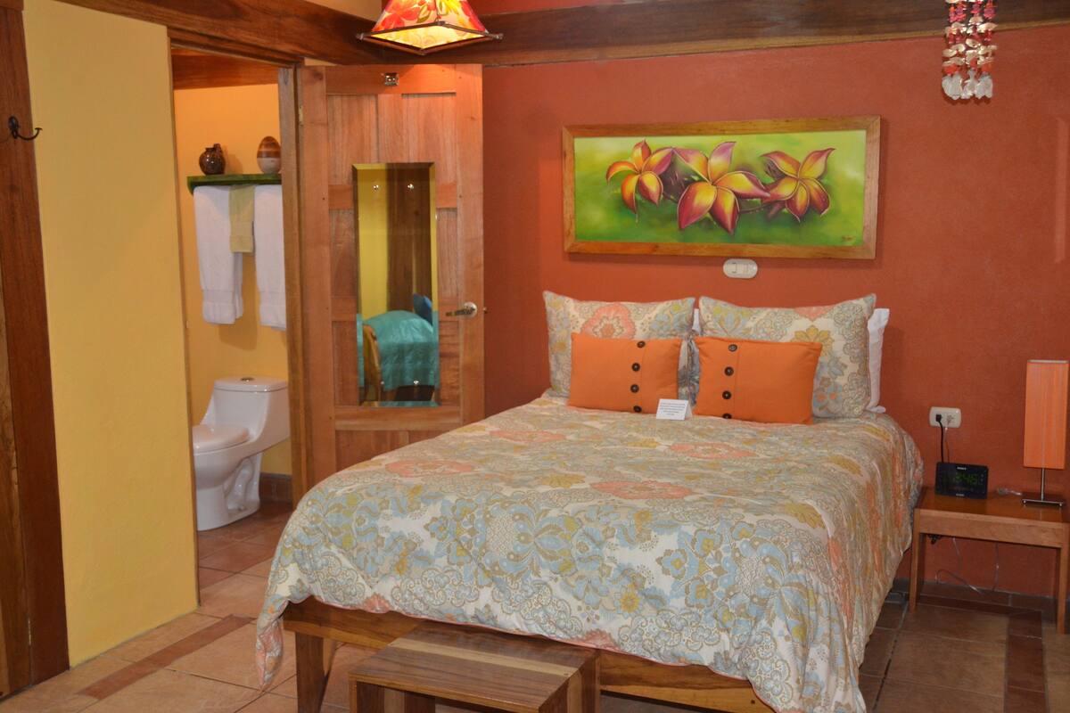 Une chambre à coucher avec un lit, une table de chevet, une commode, un tableau, une salle de bain visible à travers une porte ouverte et un luminaire suspendu.