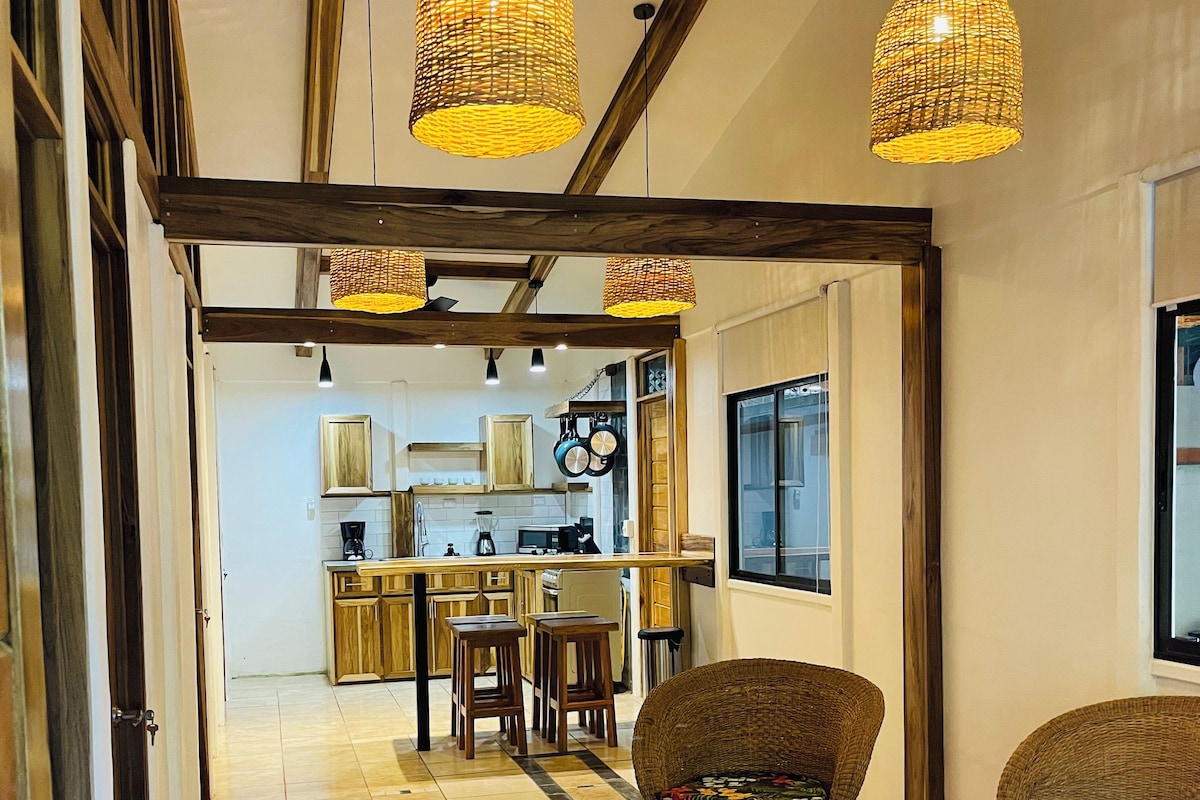 A spacious living area with a high ceiling, exposed wooden beams, and a kitchen in the background. There are wicker chairs and a dining table with stools.