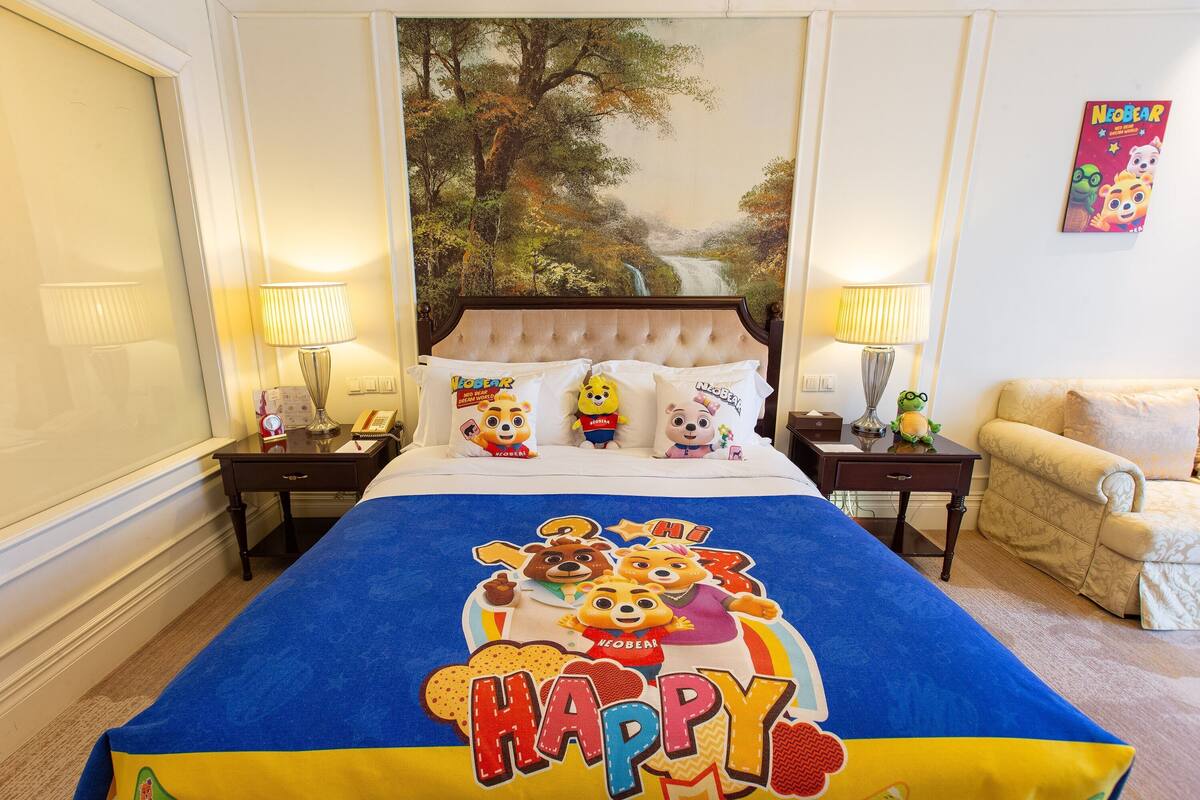 A hotel room with a large bed featuring a colorful bedspread with cartoon characters and the word "HAPPY". There are two bedside tables with lamps, a small sofa, and a wall painting of a forest scene.