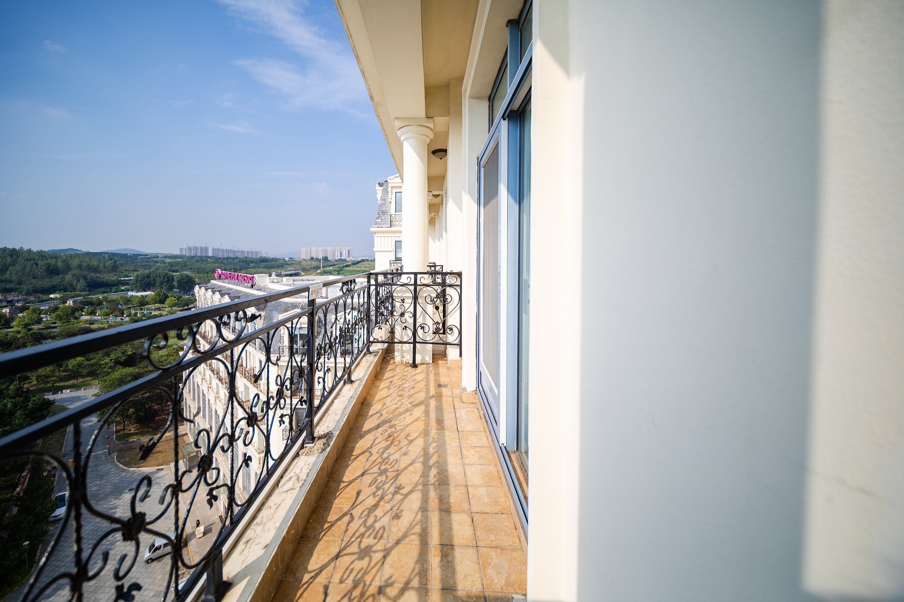 A balcony with a wrought iron railing, a tiled floor, and a view of a cityscape with buildings and greenery.