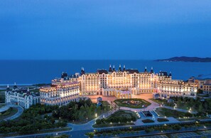 A large, multi-story hotel with a central courtyard, surrounded by greenery and a view of the sea.