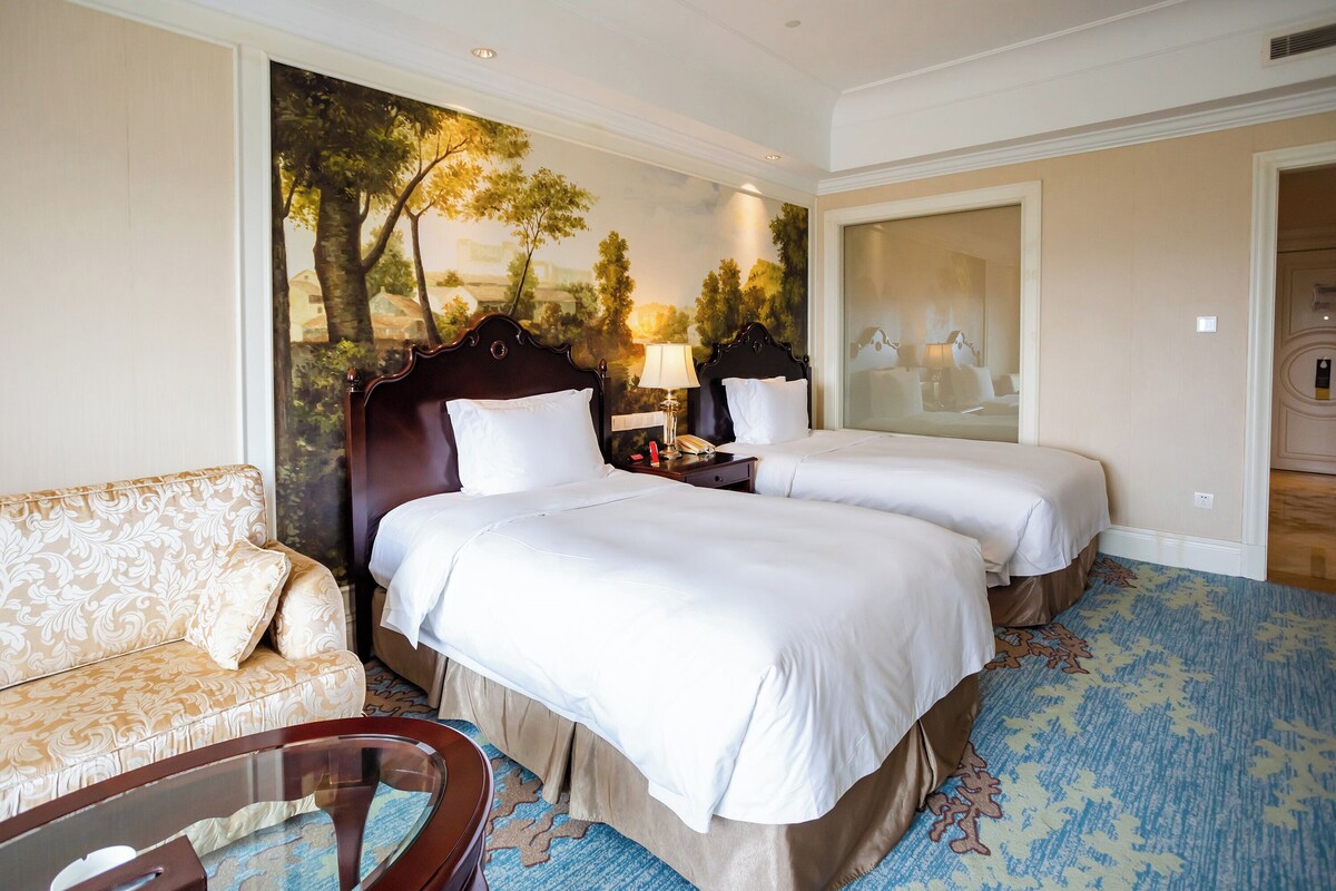 A hotel room with two beds, a sofa, a glass coffee table, and a large window with a view of trees.