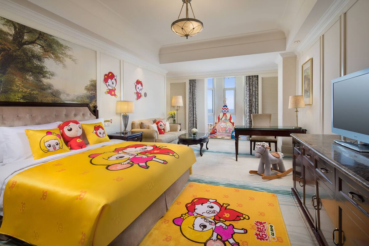 A hotel room with a large bed, a TV, a desk, and a chair. There is a painting on the wall and a window with curtains.