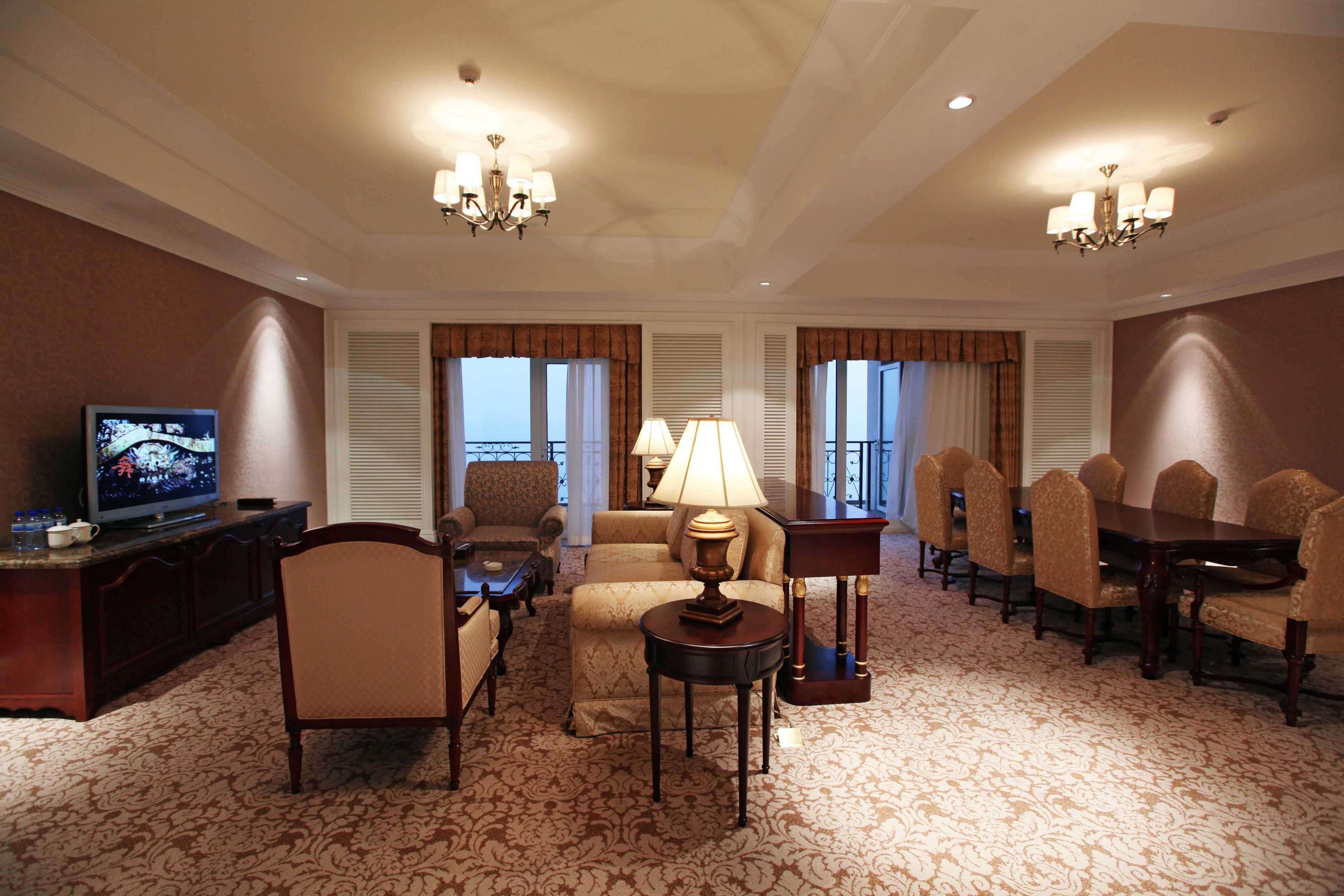 A hotel room with a television, seating area, dining table, and two chandeliers.