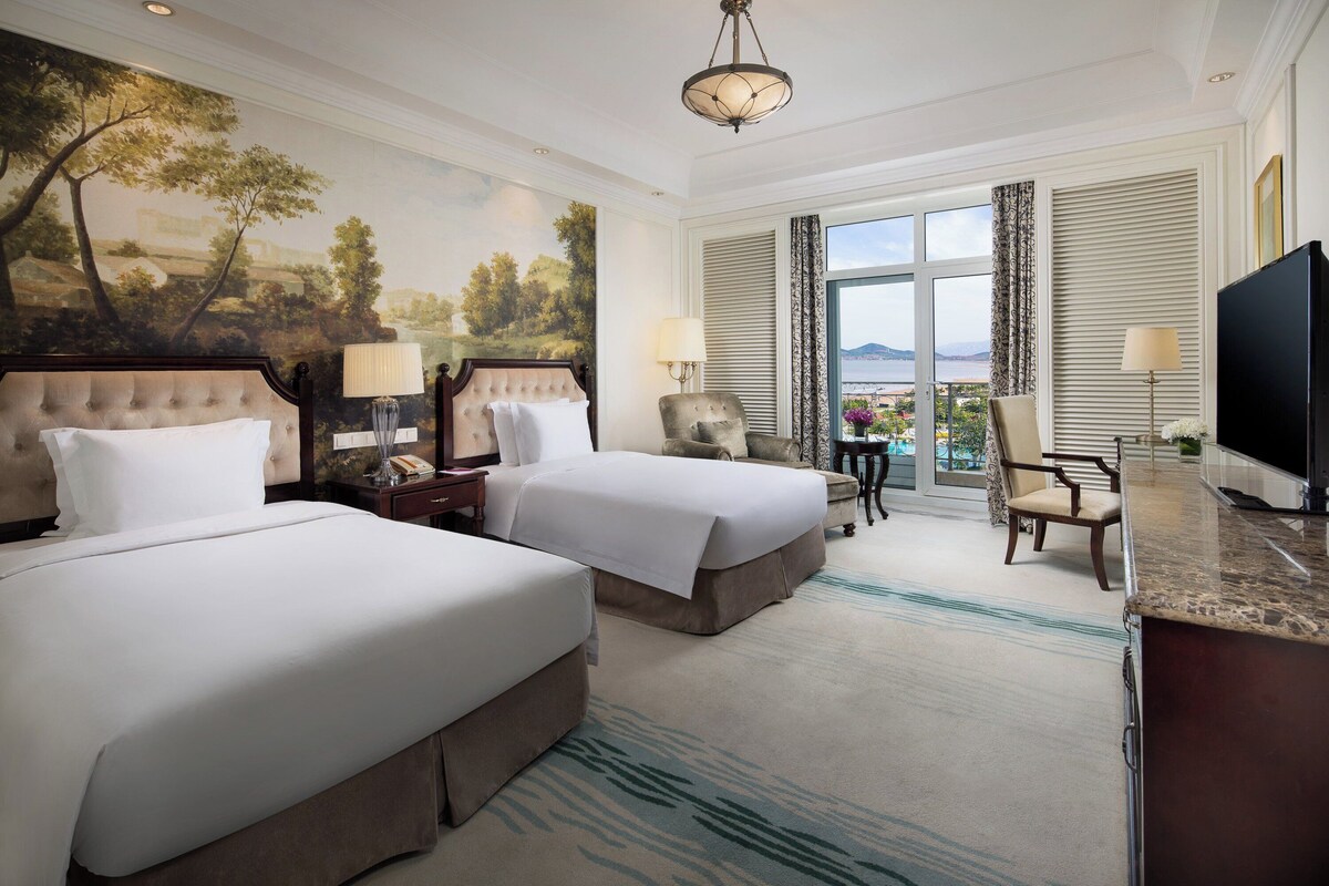 A hotel room with two beds, a sitting area, a television, and a view of the sea.