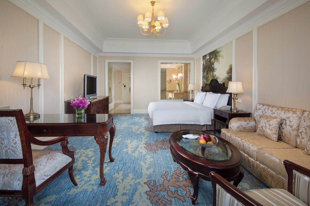 A hotel room with a bed, a sitting area with a sofa and armchair, a desk with a television, and a chandelier.