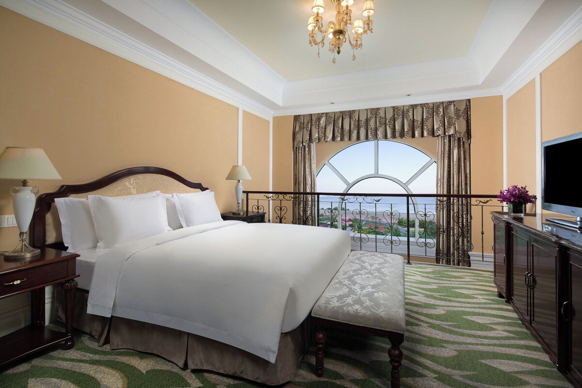 A hotel room with a large bed, a bench, a TV, a balcony with a view, and a chandelier.