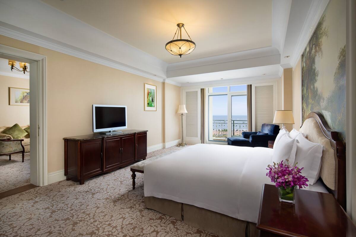 A hotel room with a large bed, a television on a wooden cabinet, a view of the outside, and a painting on the wall.