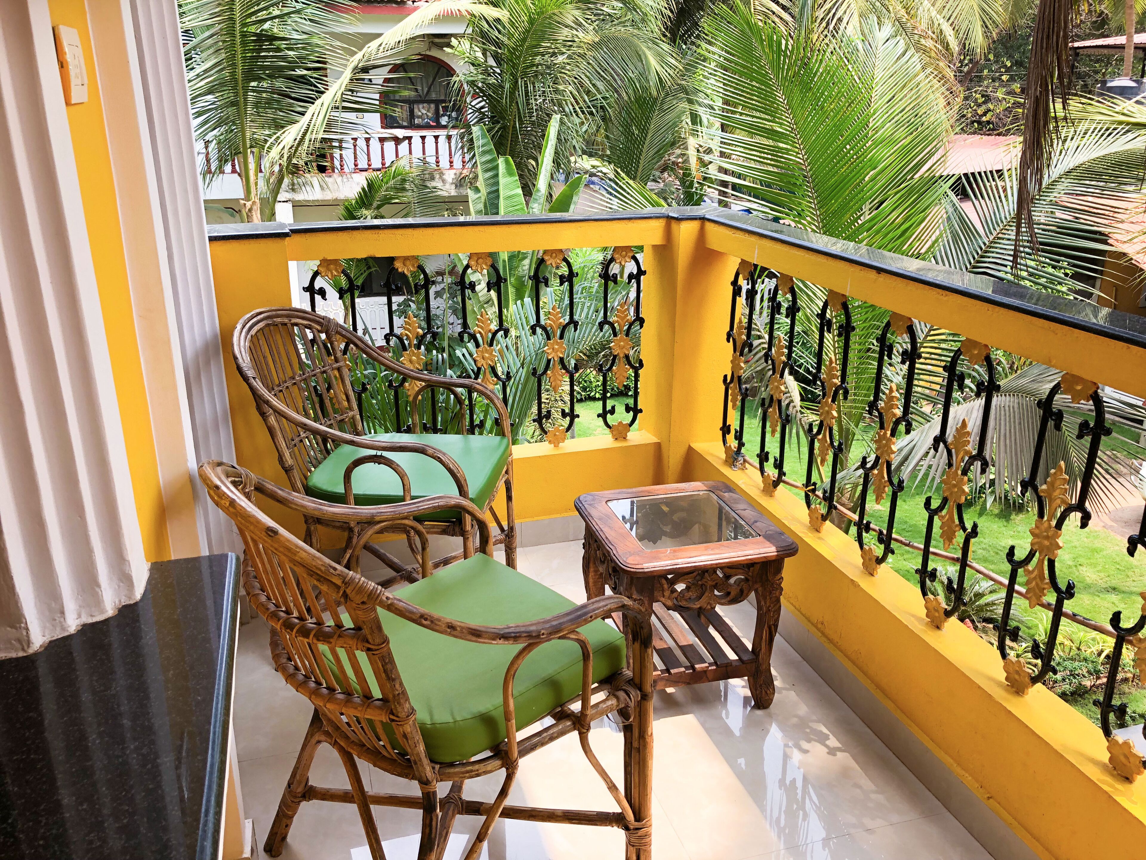 Een balkon met twee rieten stoelen en een tafeltje, gele wanden en uitzicht op een tuin met palmbomen.