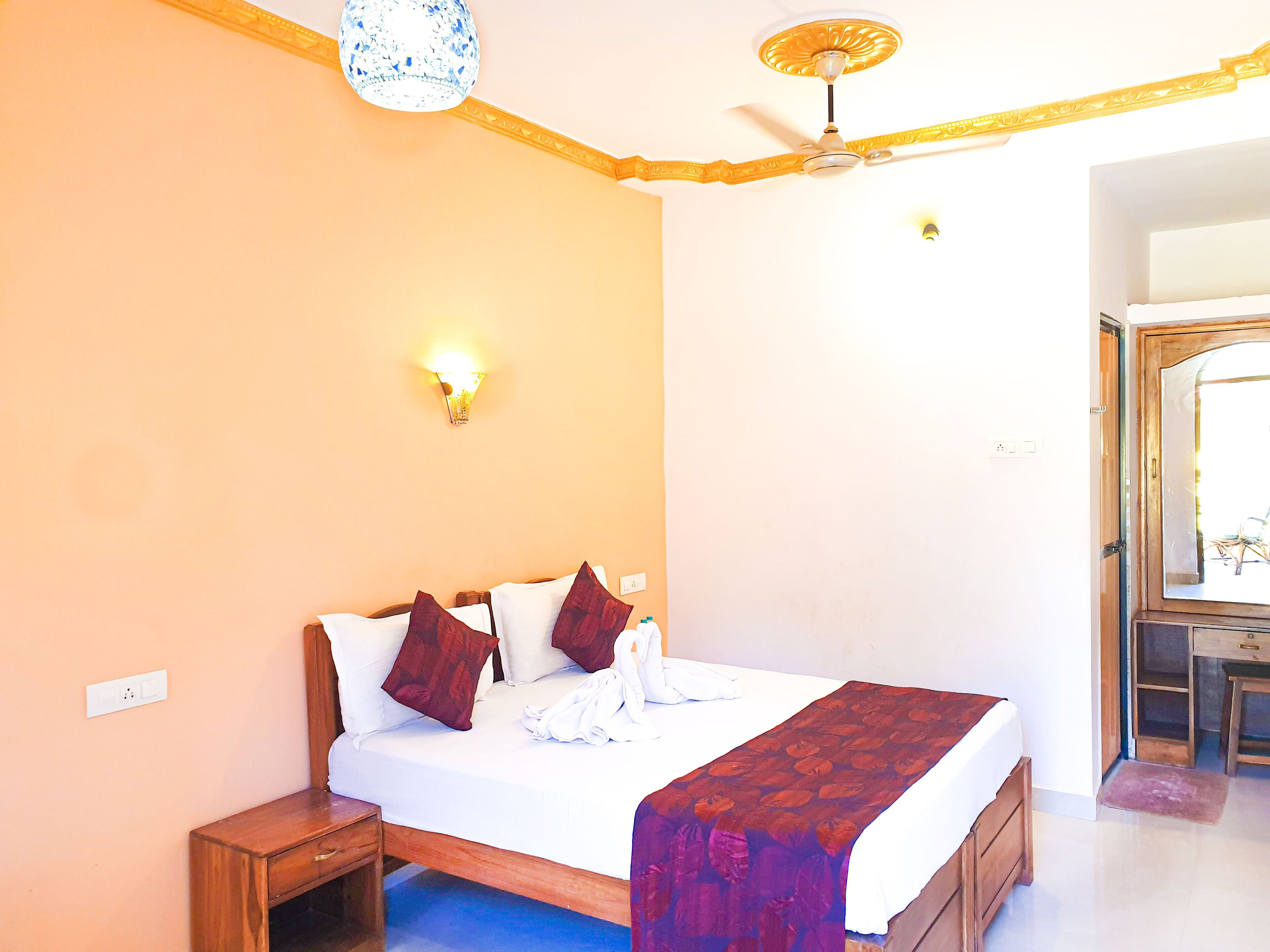 A bedroom with a bed, bedside table, wall-mounted light, ceiling fan, and a door leading to another room.