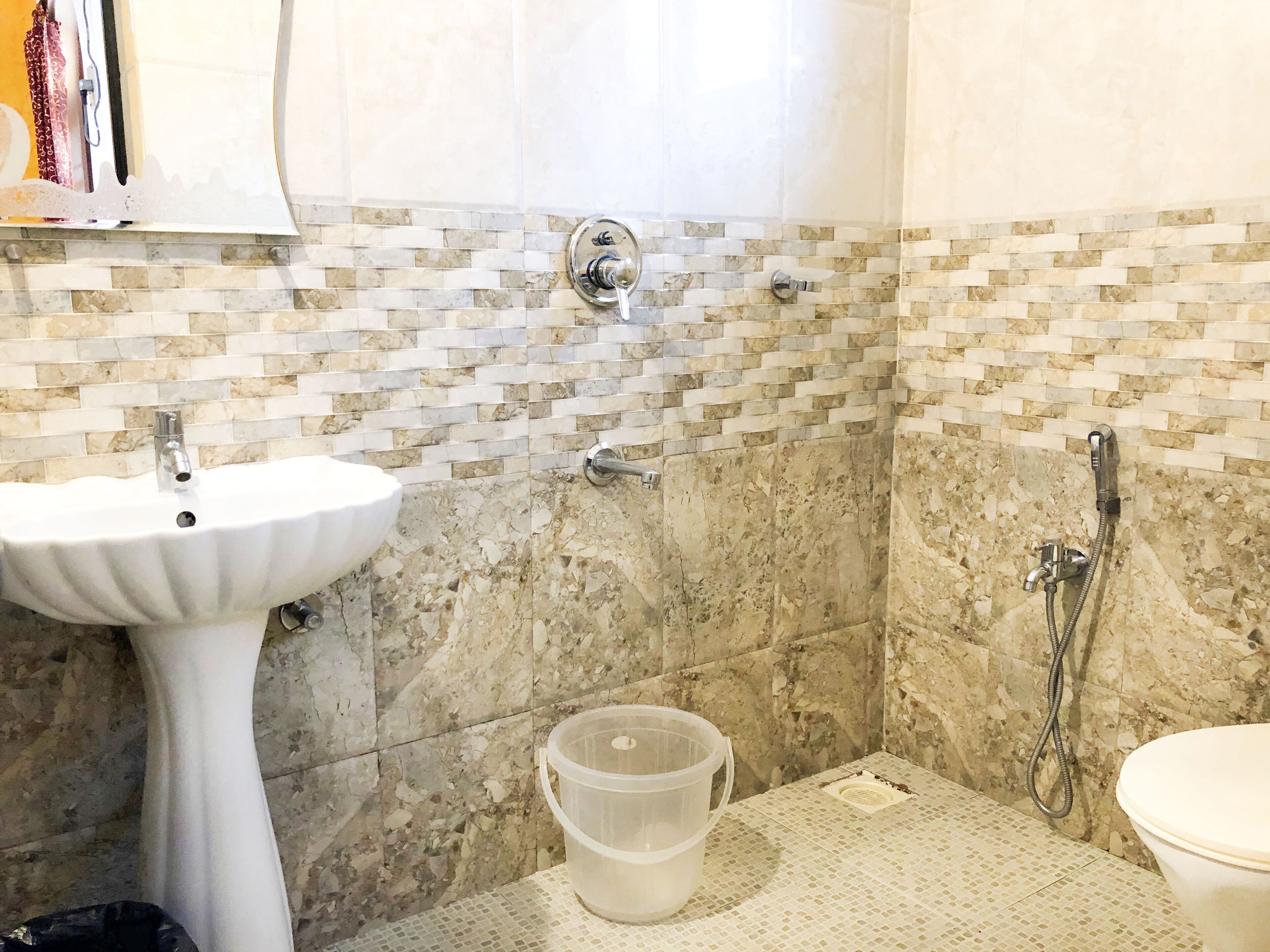 A bathroom with a white pedestal sink, a shower with a handheld showerhead, and a toilet. The walls are tiled with a mosaic pattern.
