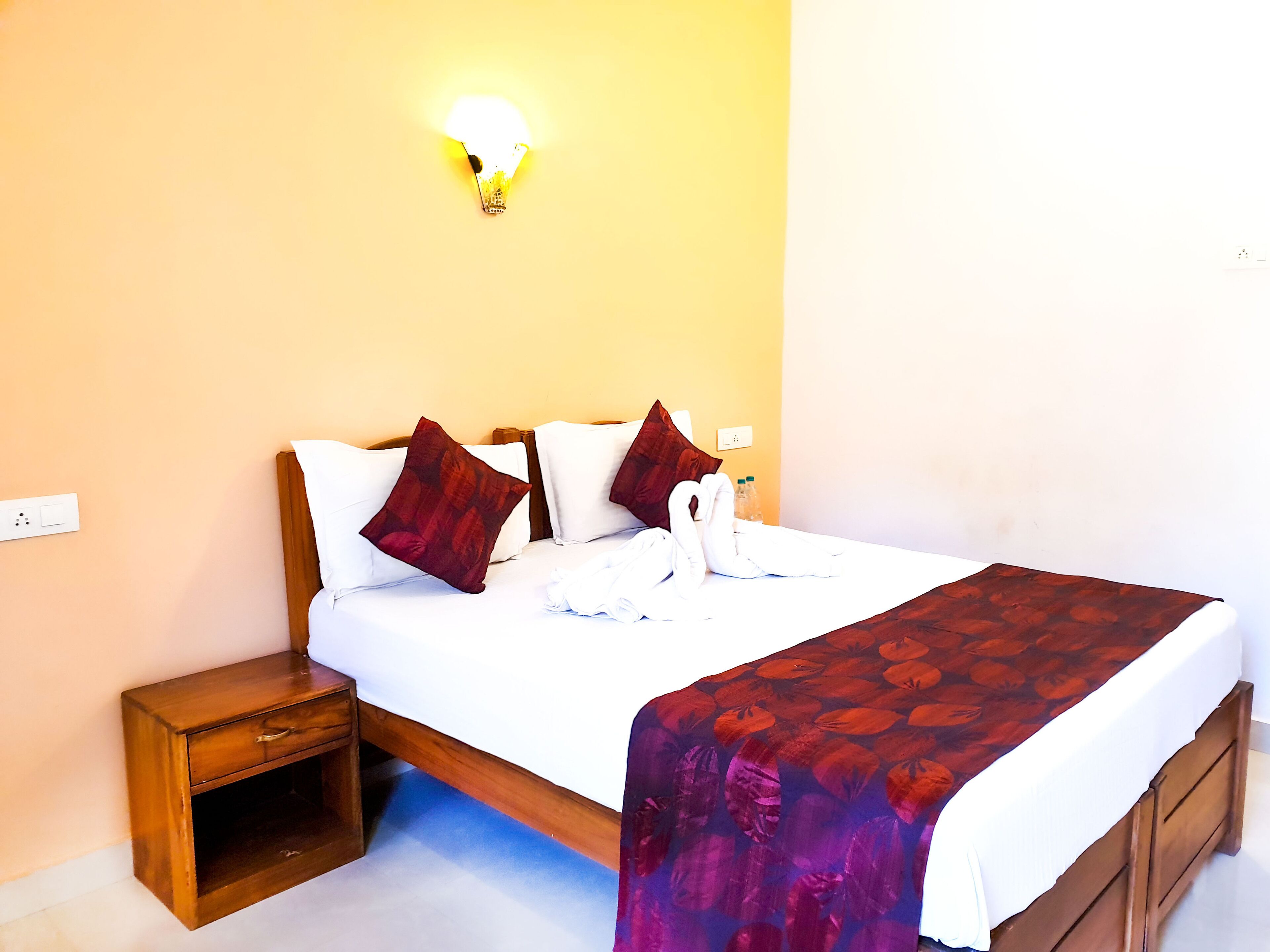 A neatly made bed with a white and red patterned bedspread, two brown pillows, and a swan-shaped towel. There is a small wooden nightstand beside the bed and a wall-mounted light fixture above it.