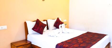 A neatly made bed with a white and red patterned bedspread, two brown pillows, and a swan-shaped towel. There is a small wooden nightstand beside the bed and a wall-mounted light fixture above it.