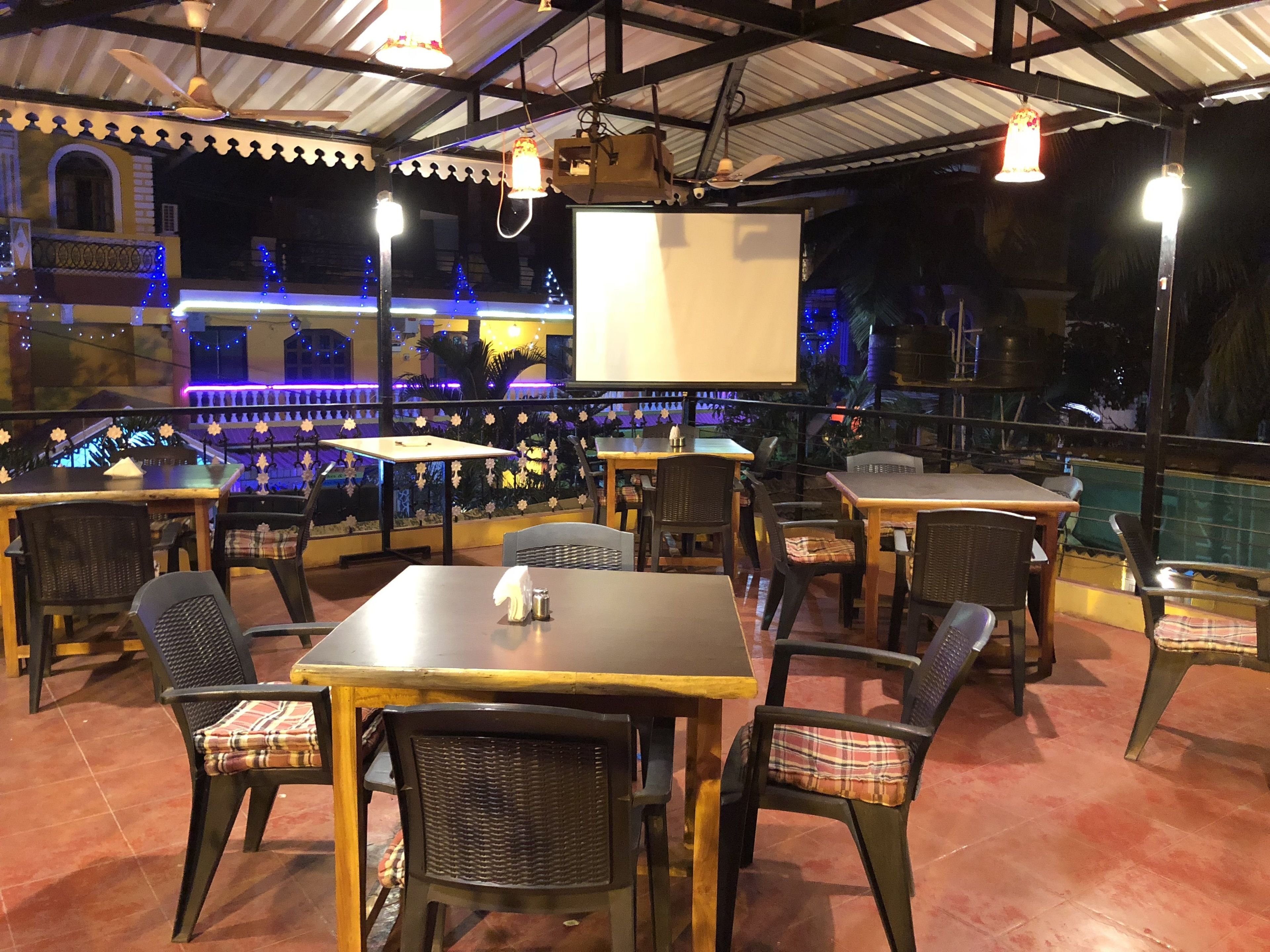 An outdoor restaurant with tables and chairs, a pool in the background, and decorative lights.