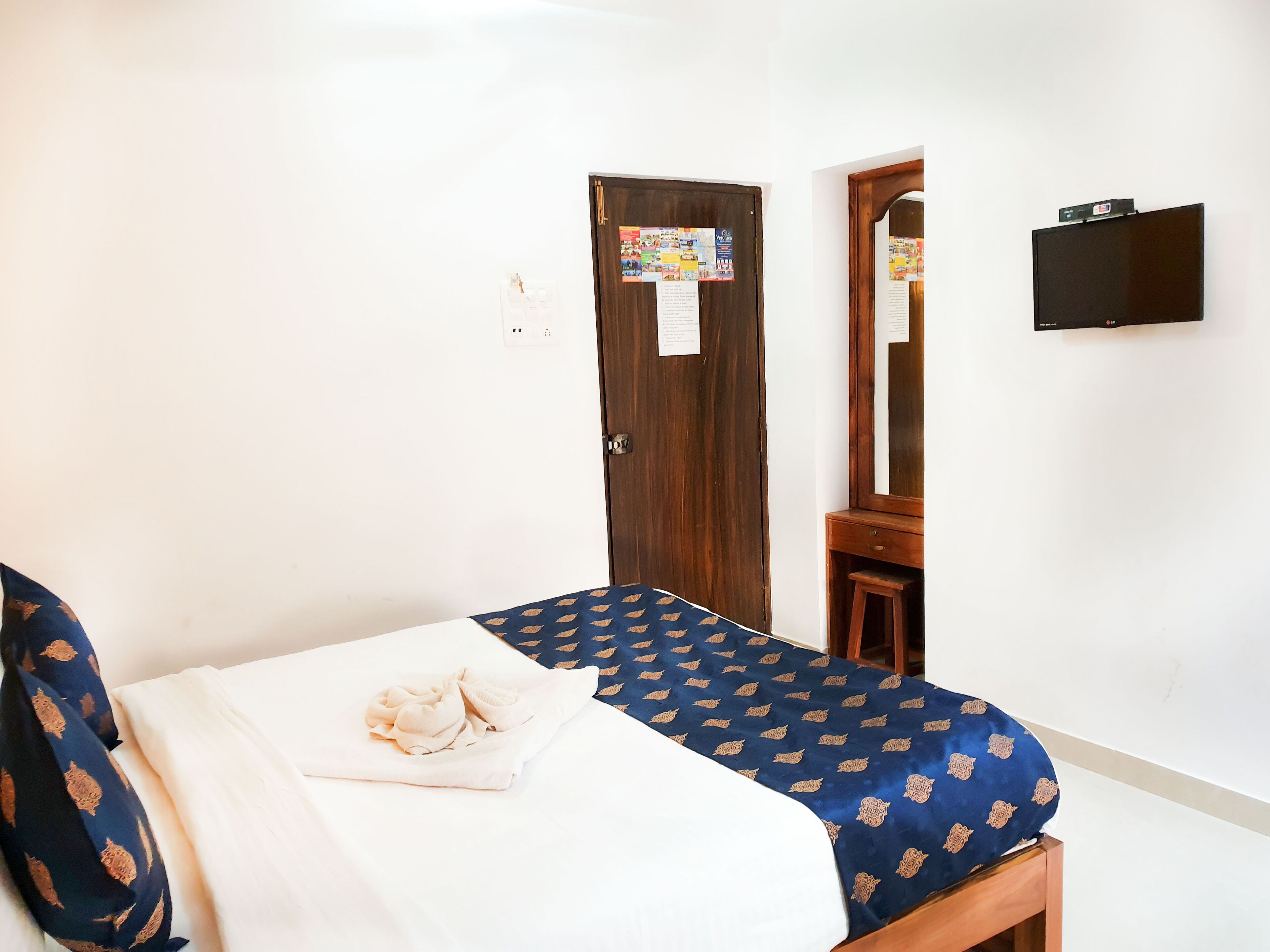 A bedroom with a bed, a wooden wardrobe, a small wooden stool, a mirror, and a wall-mounted TV.