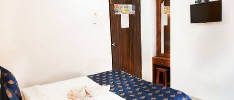 A bedroom with a bed, a wooden wardrobe, a small wooden stool, a mirror, and a wall-mounted TV.
