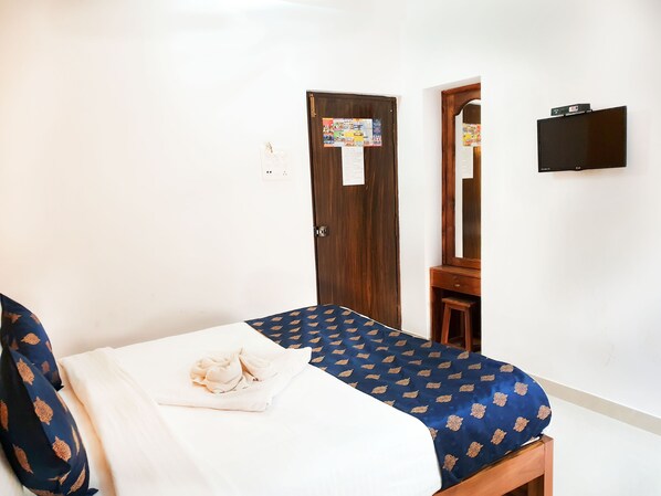 A bedroom with a bed, a wooden wardrobe, a small wooden stool, a mirror, and a wall-mounted TV.