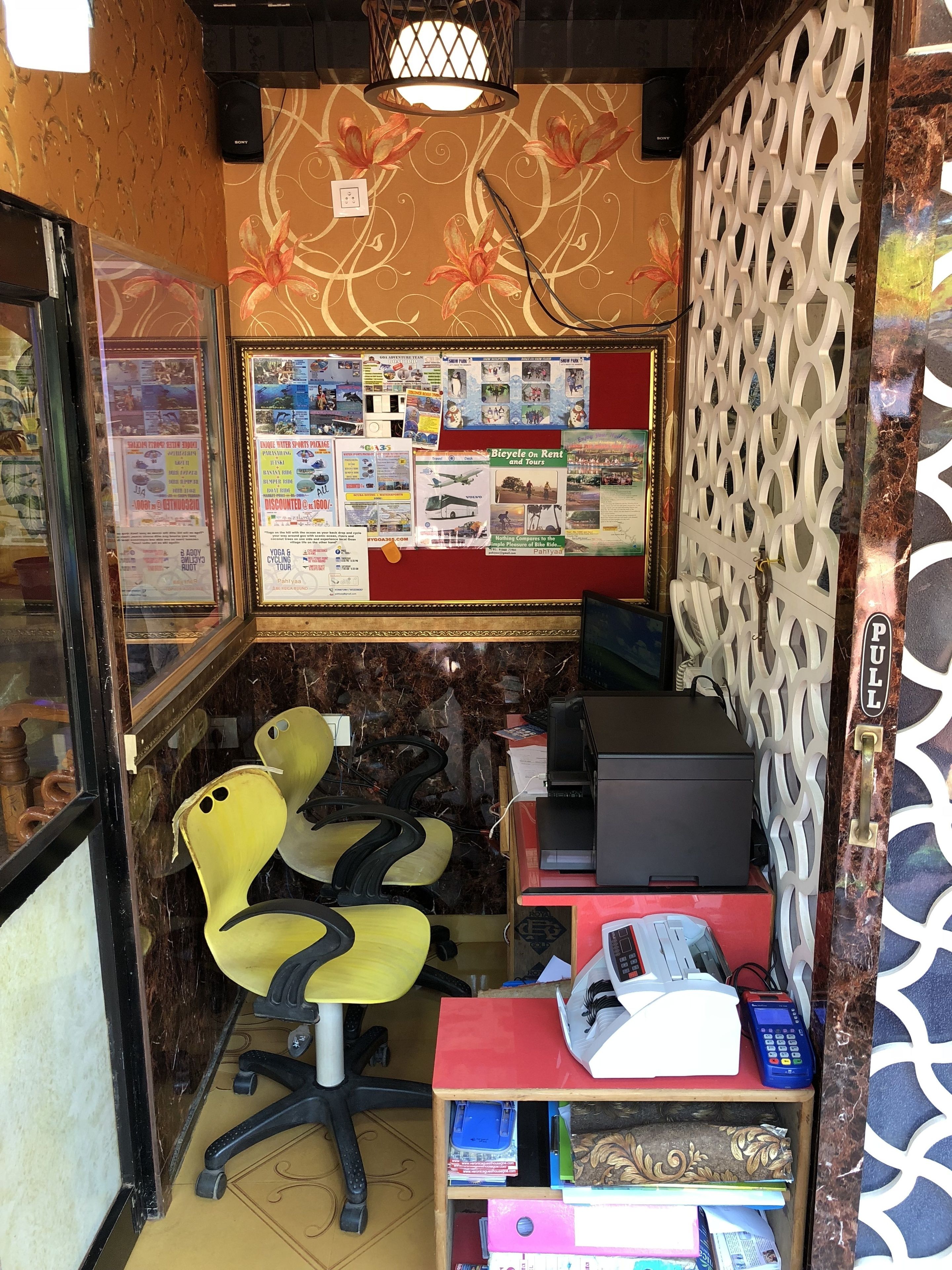 A small office with a bulletin board displaying various notices and a desk with a computer, printer, and phone.