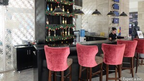 Een moderne bar met een bartender, rode fluweel barstoelen, een kerstversierde plank met flessen en een groot televisiescherm.