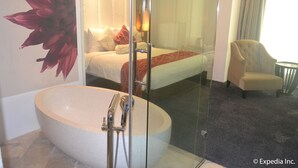 Een moderne hotelbadkamer met een groot vrijstaand bad, een douchecabine met glazen wanden, en een bed met een rode accentdeken.