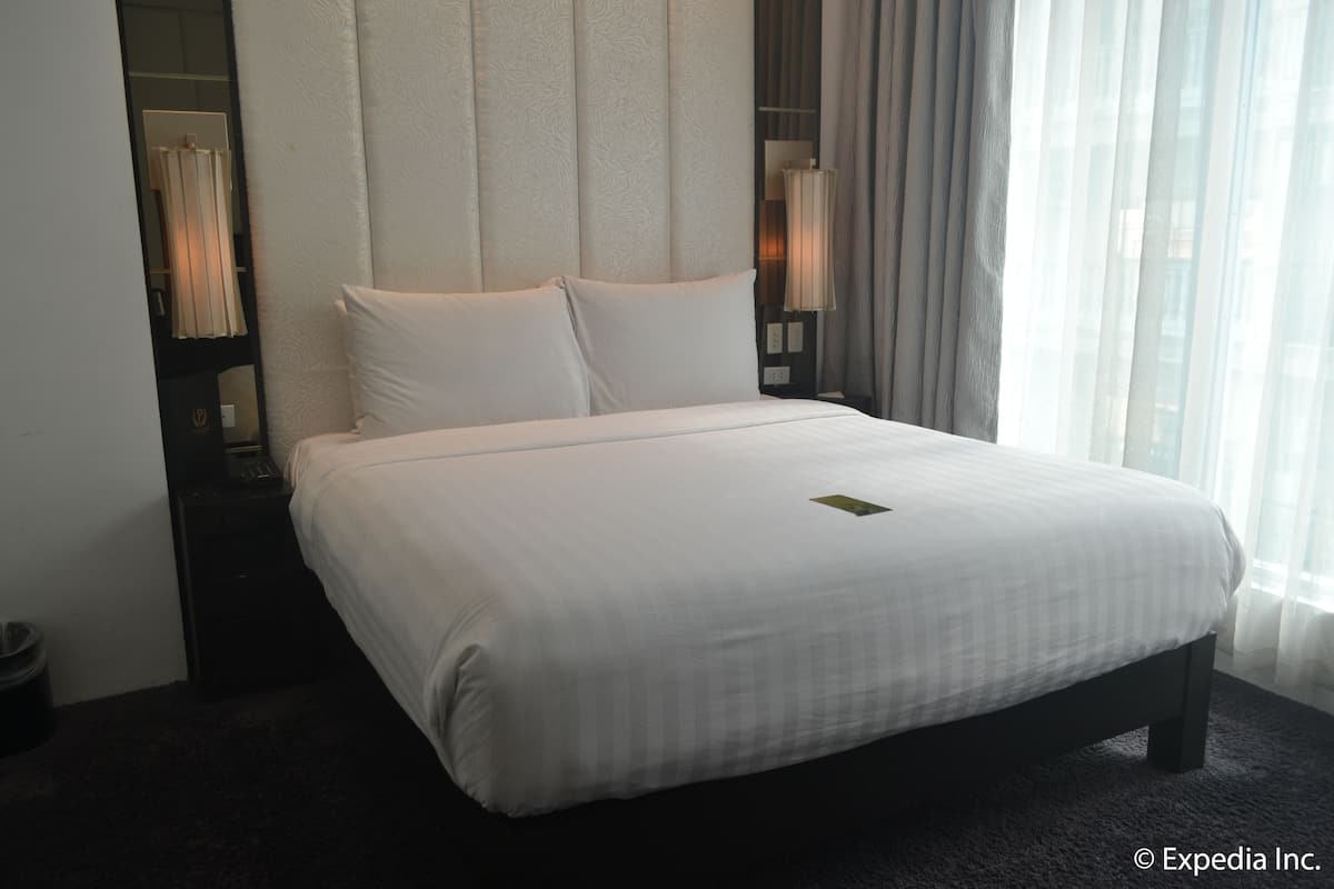 A hotel room with a large bed, a bedside table with a lamp, and a view of the city through sheer curtains.